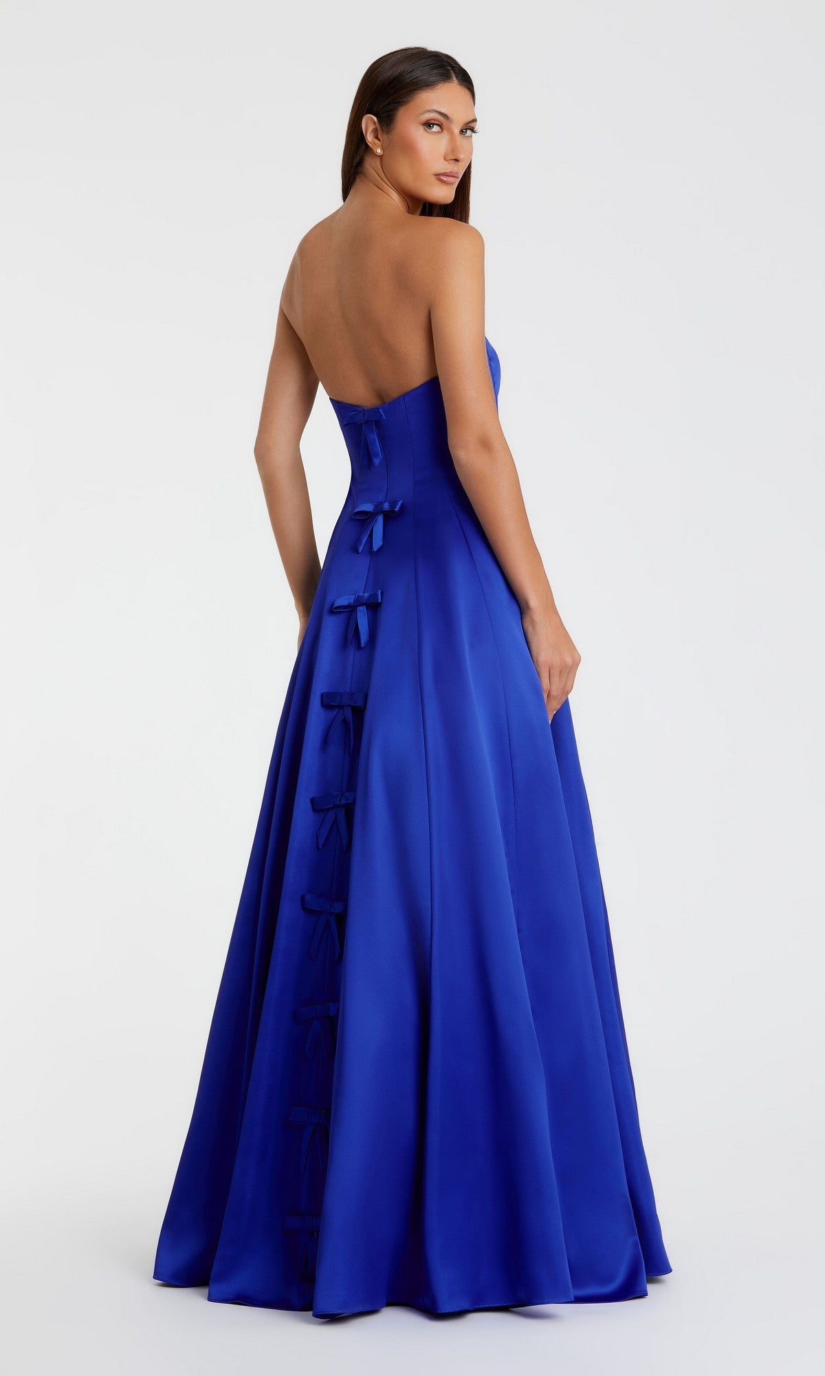 A woman models the Mac Duggal 68857 Long Formal Dress—a strapless satin gown with a fitted bodice and bow accents down the back—set against a plain light background.